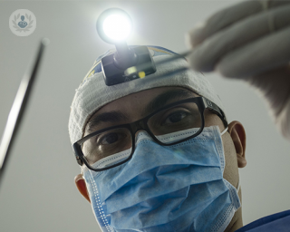 The angle suggests that a patient is lying down and about to have a procedure. The patient's view is the face of a dentist looking over them and into their mouth. The dentist is wearing a mask and holding dental tools.