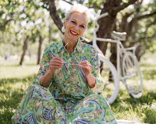 A smiling elderly woman wearing a green dress. She is sat on the grass, holding her sunglasses off her face