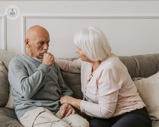 An elderly married couple sat on a sofa. The elderly man is coughing into his fist whilst the woman looks worriedly.