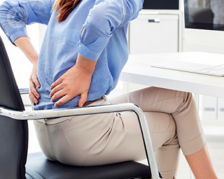A woman sat on a desk chair, holding both of her hands to her lower back, feeling pain.