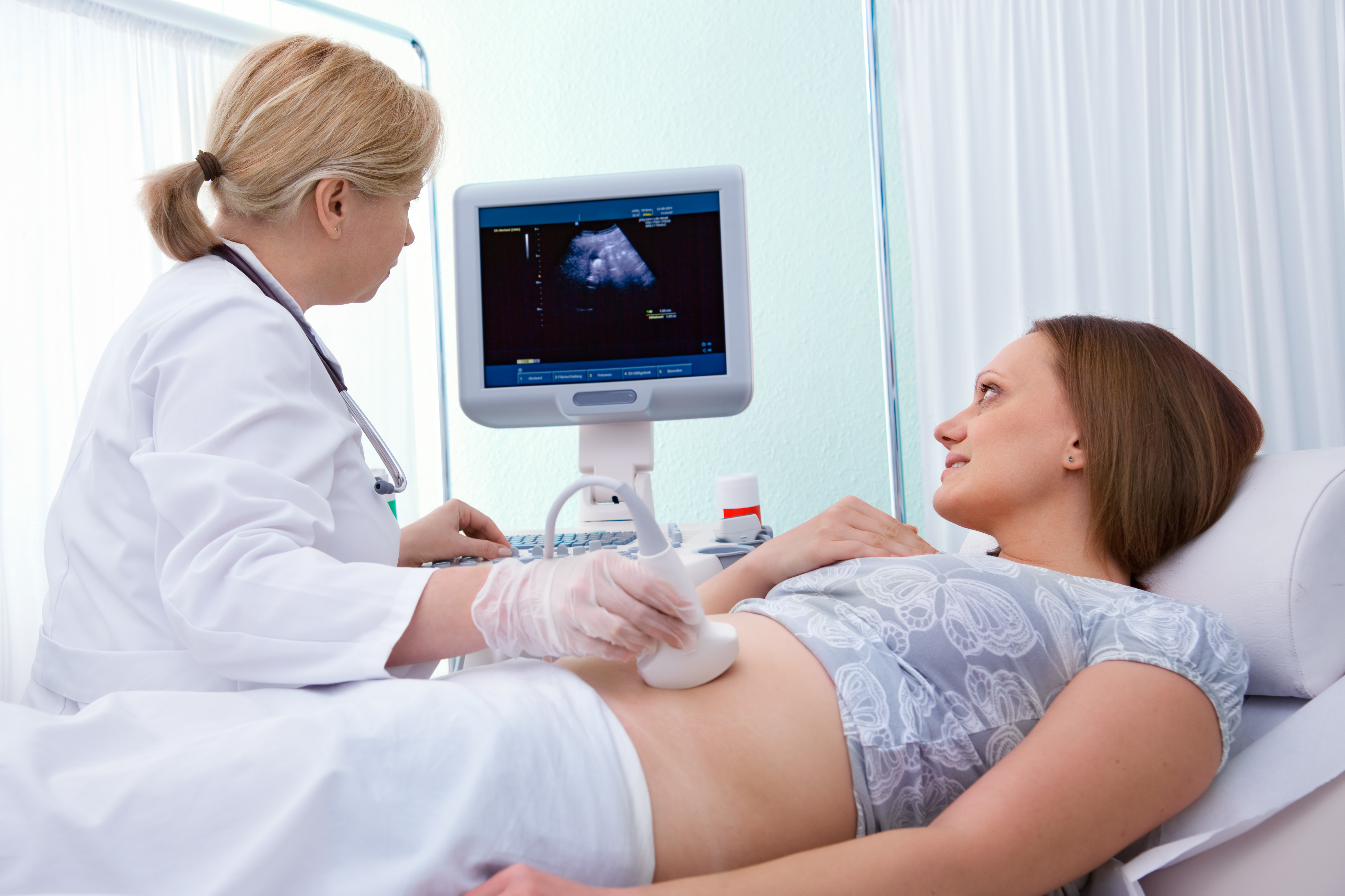 A medical specialist performing an ultrasound on a female patient. The doctor is holding the probe over the patients midsection and both patient and doctor are looking at the ultrasound imaging on a screen.