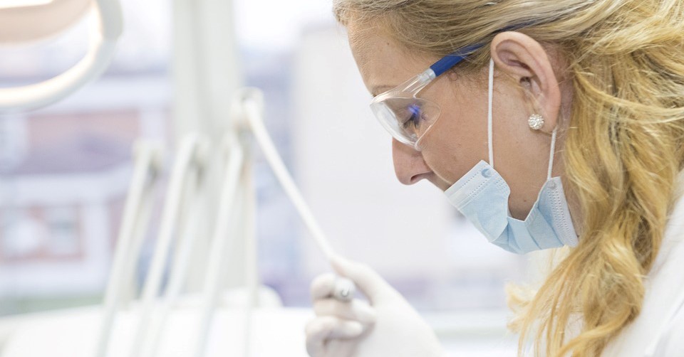 A female dentist in the middle of performing a dental procedure. She is looking down towards a patient, wearing a face mask and holding a dental tool. Cosmetic dentistry is one of the most well known specialties of Harley Street.