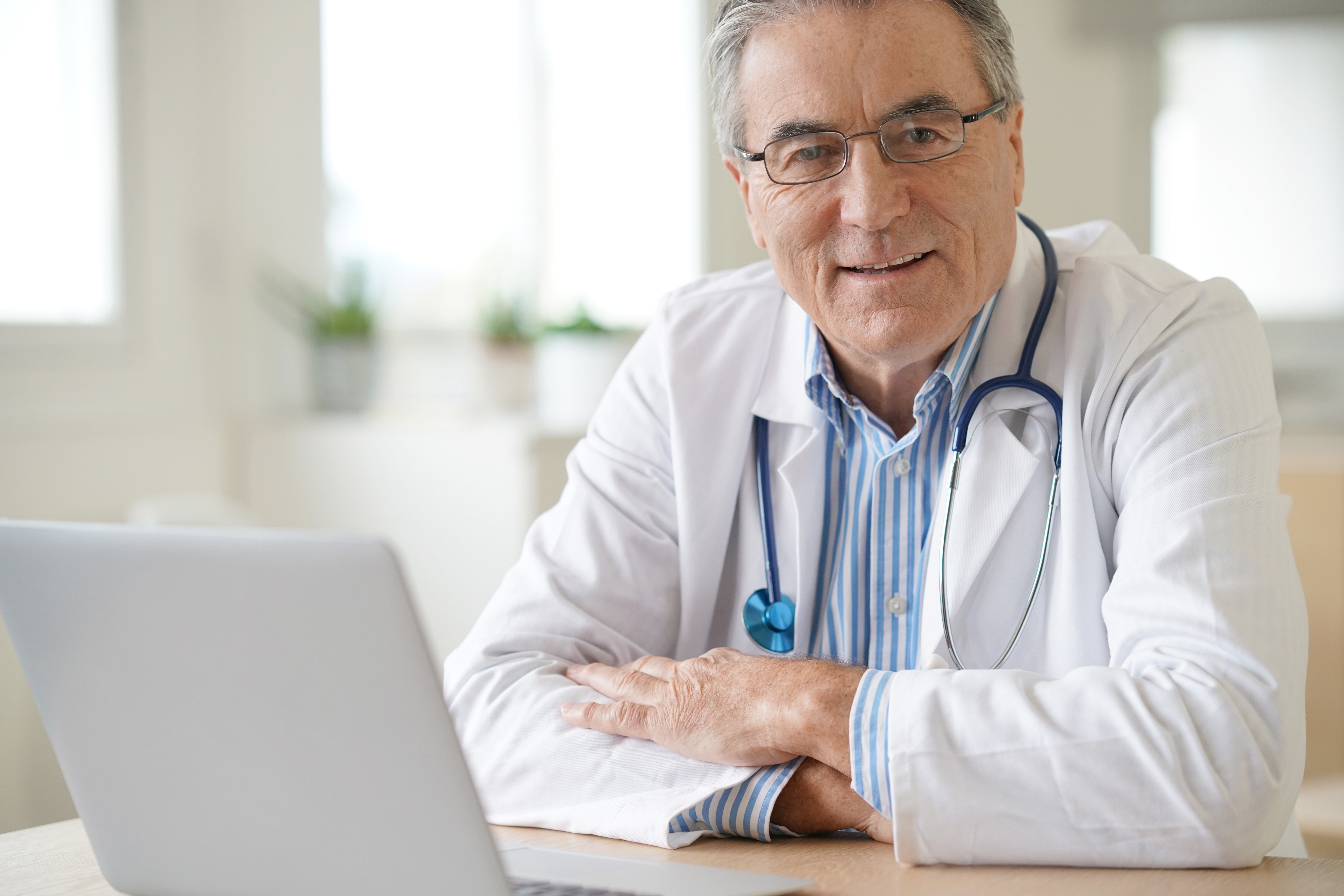 A medical specialist is sitting at a table with his arms folding over it. He's smiling and looking towards the viewer of the image. There is a laptop in front of him, which he can use for a Top Doctors e-Consultation.