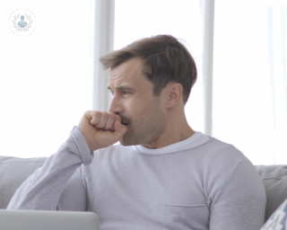 A man is sitting on his couch and coughing into his hand.