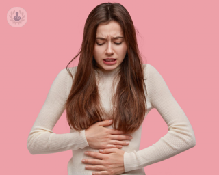 Girl holding both hands to her stomach due to acid reflux, stood against a pink background.