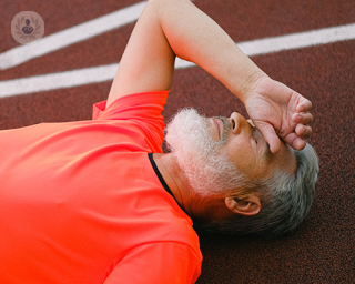 An elderly man laying down on a running track trying to catch his breath and holding a hand to his forehead