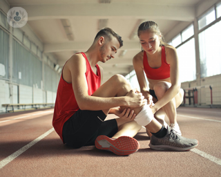 A man with a sports injury is sat on the gym floor being attended by a woman.