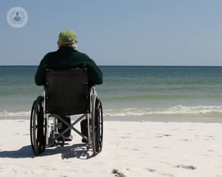 An elderly man sat on a wheelchair at a beach looking out into the sea in front of him.