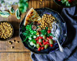 A bowl of food with chickpeas, pita bread, beans, cucumber, olives, pepper, yoghurt.