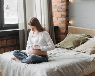 A pregnant woman sitting on her bed holding her hands to her belly