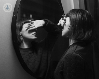 A young woman in front of a mirror, hiding her face behind her hand.