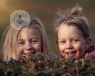 Two children are looking at the photographer. They are smiling and playful, hiding behind some grass.