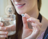 A woman's face from below her eyes is visible. She is holding a glass of water and a capsule endoscopy pill cam in front of her face.