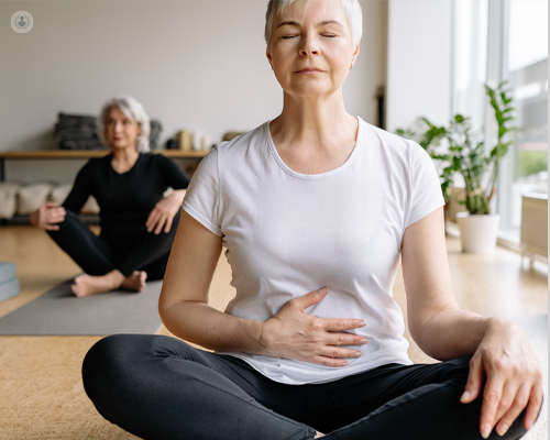Older woman calmly sat on a yoga mat with closed eyes and her hands on her stomach