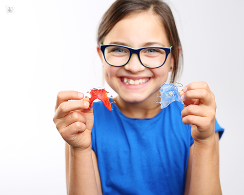 A young girl about the age of 8 years old is holding two small apparatuses in front of her. These apparatuses are braces that are placed inside the mouth behind the teeth in order to prevent and/or correct future teeth and jaw issues.