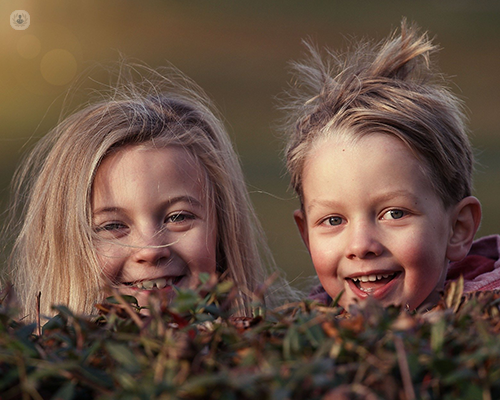 Two children are looking at the photographer. They are smiling and playful, hiding behind some grass.
