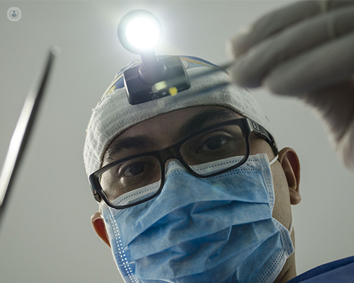 The angle of the image suggests that a patient is lying down and about to have a procedure. The patient's view is the face of a dentist looking over them and into their mouth. The dentist is wearing a mask and holding dental tools.