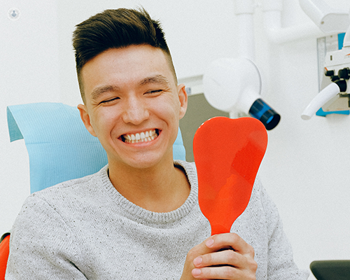 A man sitting in a dentist chair while looking at his new smile with a handheld mirror