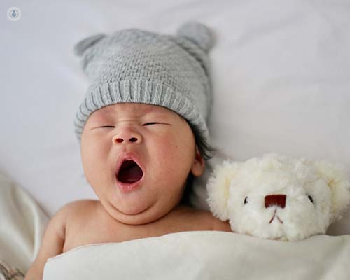 Baby yawning in cot, wearing a hat.